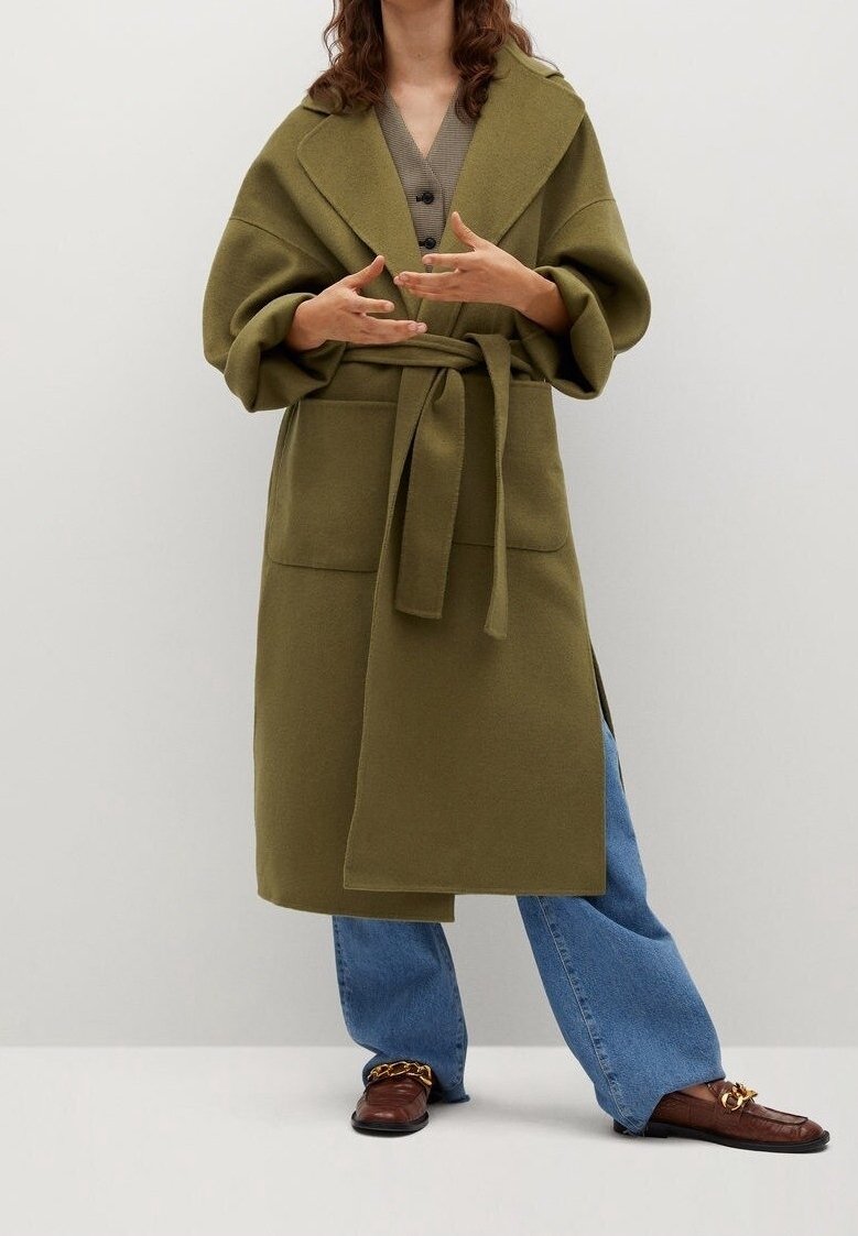 Abrigo cruzado verde oliva con cuello grande y cinturón, con grandes bolsillos delanteros, llevado sobre una camiseta gris y jeans azules, combinado con zapatos marrones.