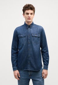 Tumši zils džinsu krekls ar pogu uz apkakles, divām krūšu kabatām ar atloka, un maigu tekstūru. Garas piedurknes ar pogām.