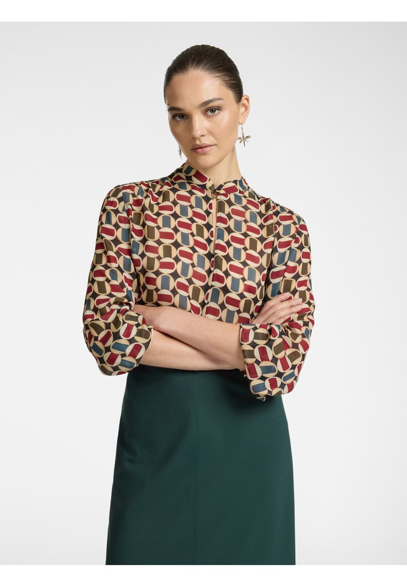 Blusa a fantasia con colletto alto, caratterizzata da forme geometriche nei colori rosso, beige e blu. Abbinata a una gonna verde, che presenta una texture liscia.