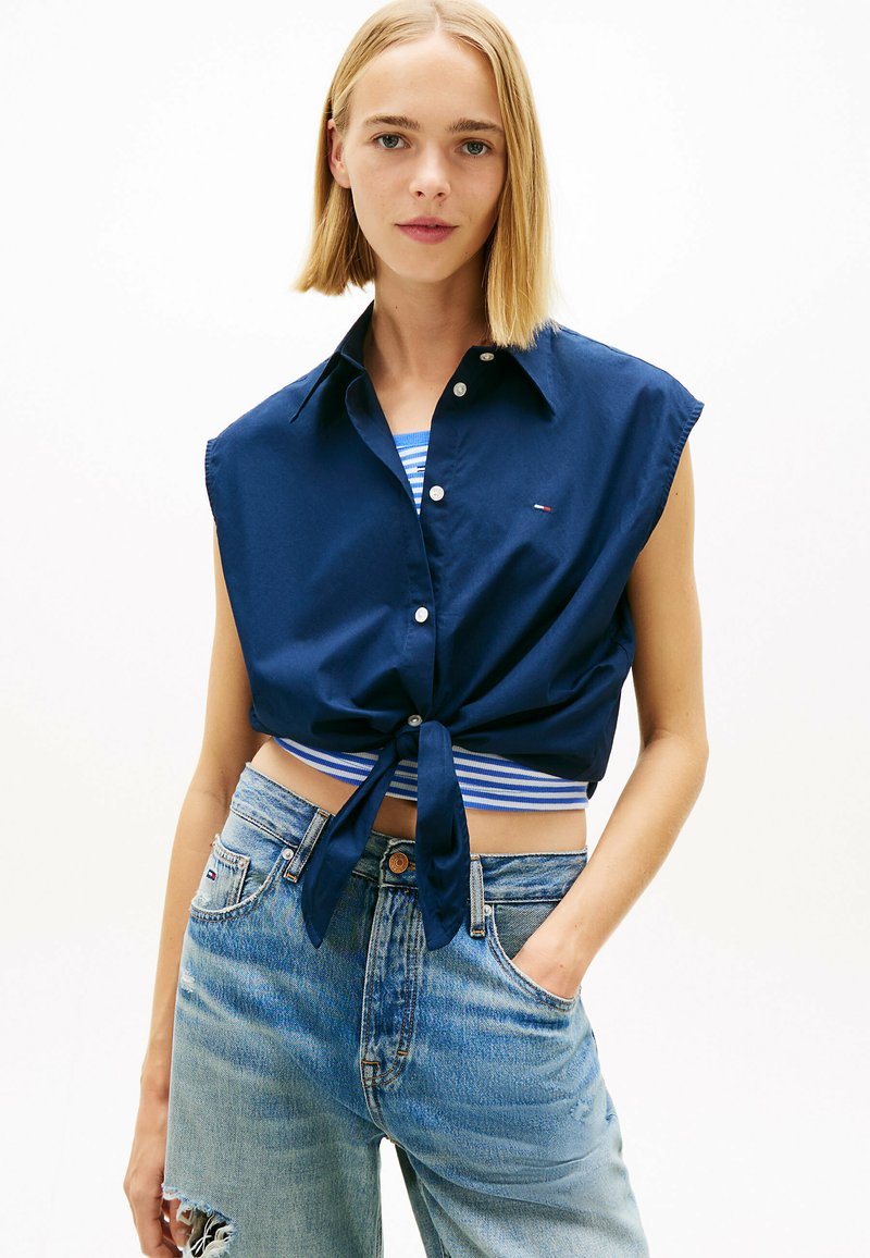 Jeune femme aux cheveux blonds coiffés en carré, portant une chemise courte bleu marine sans manches boutonnée et nouée à la taille, un haut rayé bleu et blanc, et un jean bleu clair déchiré.