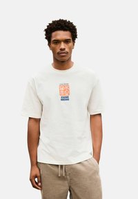 Rövid, göndör hajú férfi, fehér pólót visel, melyen narancssárga és kék felirattal az "Utagawa Hiroshige" szerepel, és bézs kordbársony nadrágot húzott, melynek derékán húzózsinór található.