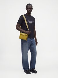 Άνδρας που φοράει μαύρο t-shirt Emporio Armani, μπλε τζιν, μαύρα παπούτσια και κίτρινη τσάντα χιαστί με φαρδιά λουράκι με λογότυπο.