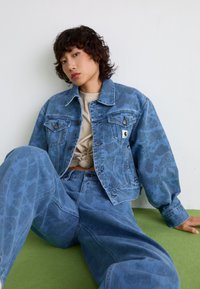 Veste en denim avec un motif camouflé bleu, coupe oversize, poches avant et petite patch logo, assortie à un pantalon en denim.