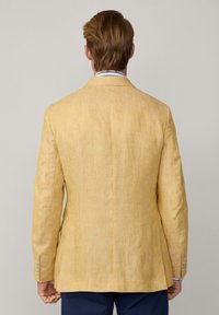 Blazer en lin jaune clair avec une fente unique, coupe ajustée et texture subtile. Présente trois boutons sur chaque poignet. Vue de dos.