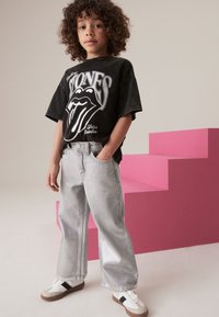 Dziecko w czarnym, oversize'owym T-shircie z grafiką Rolling Stones, jasnoszarej szerokiej parze dżinsów i białych sneakersach z brązowymi akcentami.
