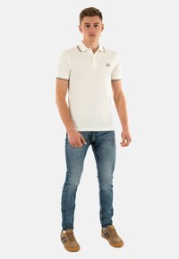 Hvid polo skjorte med en to-knaps knaplukning, stribet krave, broderet logo. Båret med lyseblå jeans og beige sneakers.