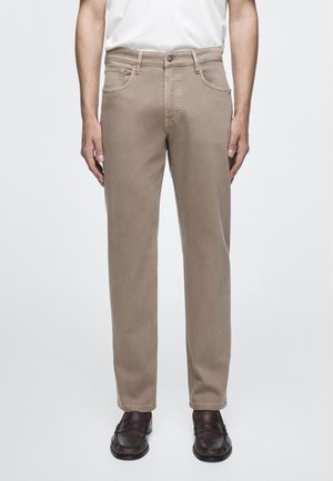 Mann trägt beige gerade geschnittene Hose, weißes Hemd eingesteckt und dunkelbraune Ledermokassins, steht vor einfachem Hintergrund.