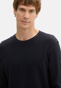 Marineblaues langärmliges Rippoberteil mit Rundhalsausschnitt; strukturierter Stoff, mit einem Logo auf dem linken Ärmel. Elegantes, lässiges Design.