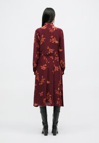 Μπορντό φόρεμα με λουλούδια, μακριά μανίκια, με κλειστό γιακά και σχέδιο Α-line. Διαθέτει σχέδια με πορτοκαλί και ροζ λουλούδια, φορεμένο με μαύρες μπότες μέχρι το γόνατο.