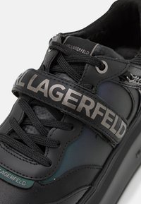 Svart sneaker med en texturerad läderövre del, tonala snören och ett framträdande grått rem med "KARL LAGERFELD"-profilering över framsidan.