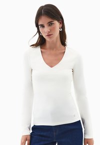 Joven mujer con cabello largo marrón, viste una blusa blanca de manga larga con escote en V y jeans azules, de pie contra un fondo blanco liso.