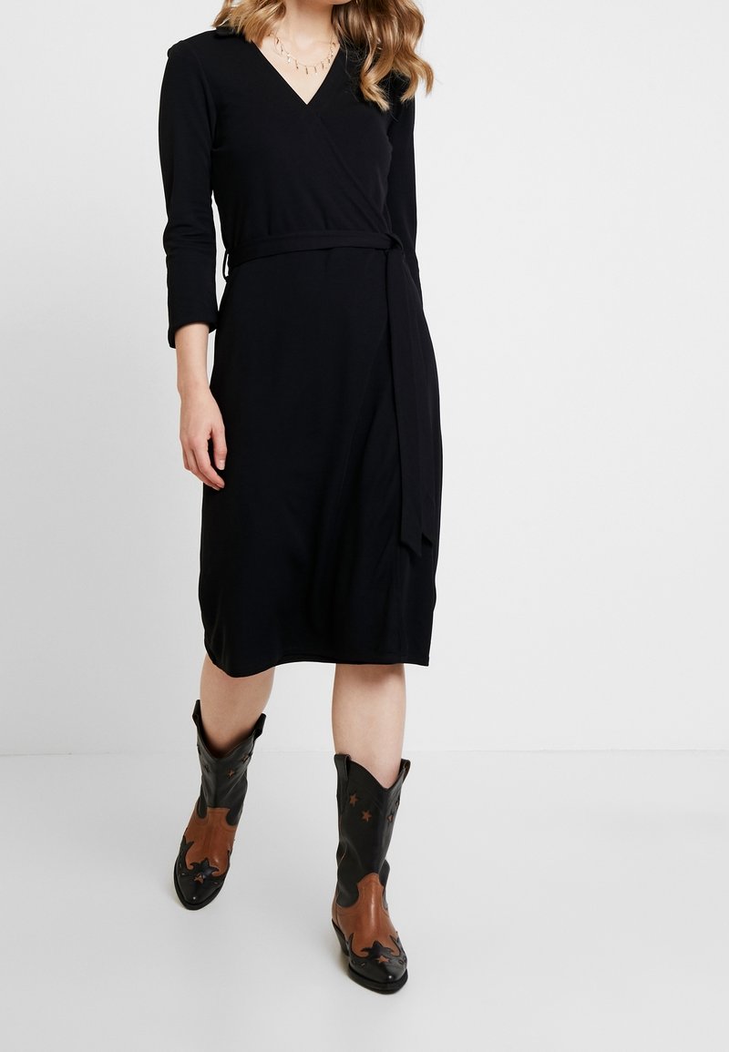 Mustapukuinen, pitkähihainen itsevaate, joka on valmistettu sileästä kankaasta ja jossa on vyötärölle solmittava nauha. Yhdistetty mustiin ja ruskeisiin cowboy-kenkään.