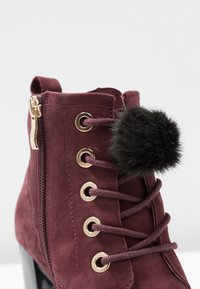 Botín de ante burdeos con herrajes dorados, diseño con cordones y un acento de pompón de piel sintética negra. Incluye un zipper lateral para facilitar su uso.