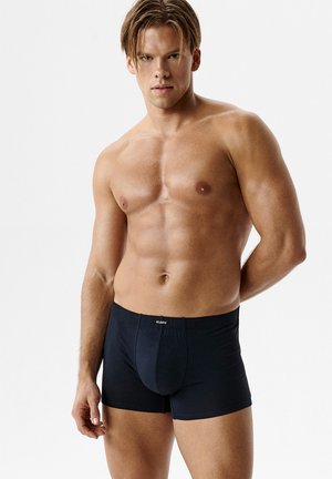 Mężczyzna bez koszulki o muskularnej budowie ciała, ubrany w czarne bokserki Atlantic, stoi na tle jednolitego białego tła.