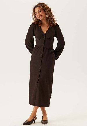 Kvinna med lockigt hår som ler, klädd i en lång svart knäppt klänning med långa ärmar och svarta slingback-klackar, stående mot en ljus bakgrund.