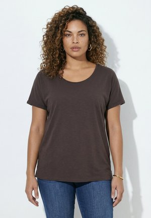 Femme aux cheveux bouclés portant des boucles d'oreilles en anneau, un t-shirt marron foncé à manches courtes, un jean bleu et un bracelet en or, debout devant un fond blanc.