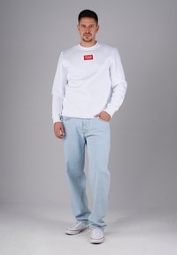 Mann steht mit einer Hand in der Tasche, trägt einen weißen Pullover mit rotem Logo, hellblaue Jeans und weiße Sneakers vor einem grauen Hintergrund.
