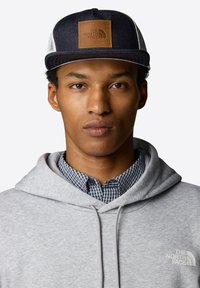 Cappellino da baseball in denim scuro con una toppa in pelle marrone con il logo "THE NORTH FACE". Felpa grigia con una camicia a quadri blu sotto.