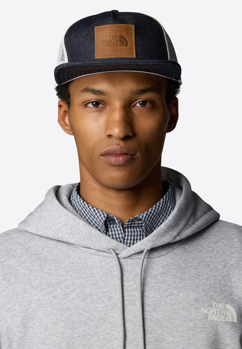 Cappellino da baseball in denim scuro con una toppa in pelle marrone con il logo "THE NORTH FACE". Felpa grigia con una camicia a quadri blu sotto.