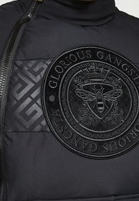 Czarna pikowana kurtka z teksturowanym wzorem tkaniny, z dużą wytłaczaną okrągłą naszywką z misternymi detalami pszczoły i korony.