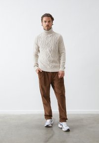 Cream knitted sweater with a high neckline and cable patterns, paired with brown corduroy pants and white athletic shoes.