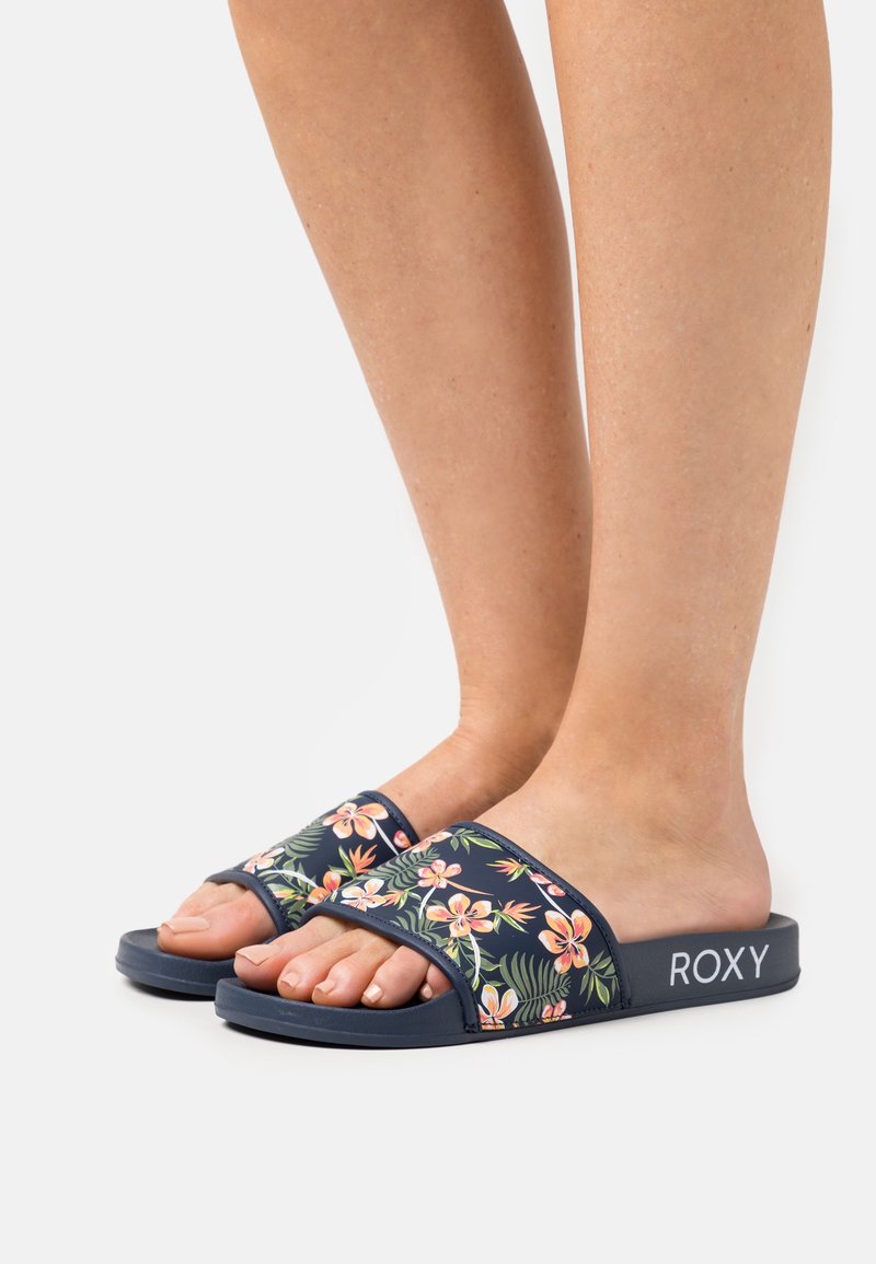 Pieds portant des sandales slides bleu marine avec des motifs floraux roses et orange et le nom de la marque ROXY sur le côté.