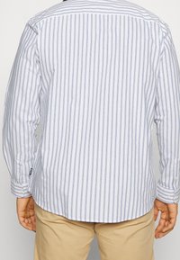 Valkoinen ja vaaleansininen, pitkähihainen raidallinen paita, jossa on nappikaulus, pyöristetty helma ja säädettävät kalvosimet. Takakuvassa näkyvät pystysuorat raidat.