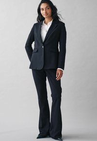 Marineblåt skræddersyet jakkesæt med blazer, der har spids revers, to lommer og en enkelt knaplukning, parret med tætsiddende bukser og spidse hæle.