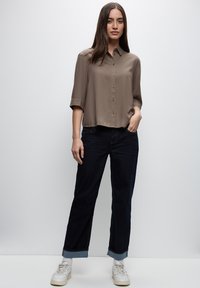 Blusa color marrone chiaro a maniche corte con collare abbinata a jeans di denim blu scuro. I jeans presentano un orlo risvoltato e sono indossati con sneaker bianche.