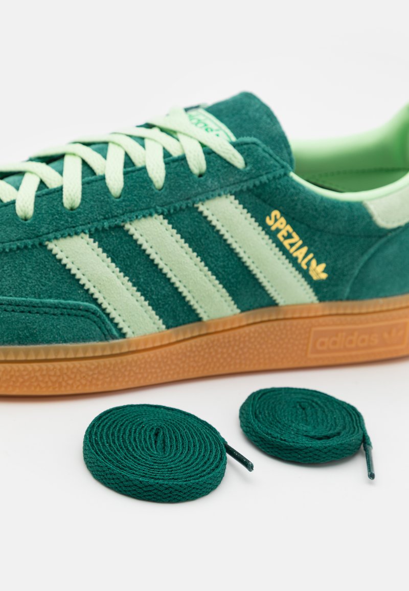 Sneaker verde in suede con strisce testurizzate bianco sporco, marchio dorato e suola in gomma marrone; include due lacci verdi arrotolati.