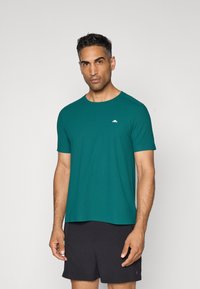 Teal kurzärmliges T-Shirt mit Rundhalsausschnitt und dezentem Logo auf der linken Brust, aus leichtem Stoff, kombiniert mit schwarzen Shorts.