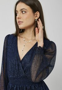 Robe bleu marine en tissu texturé, décolleté en V profonds, et manches bouffantes transparentes. Les accessoires comprennent un collier en argent avec une lasso et des boucles d'oreilles créoles.