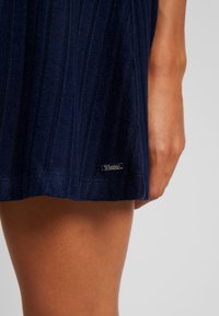 Tmavě modré žebrované pletené šaty s metalicky stříbrnou logem na lemu. Tkanina má strukturovaný povrch a rozšířený tvar.