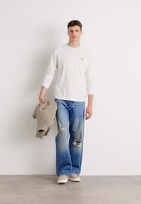 Witte blouse met lange mouwen en een klein logo, gecombineerd met lichtblauwe relaxed-fit versleten jeans. Beige jas in één hand, neutrale schoenen.