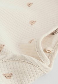 Tissu crémeux à côtes avec des imprimés d'ours brun clair, présentant une texture douce, des bords arrondis et un col replié.