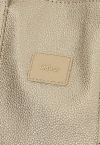 Sac à main en cuir texturé doré avec une étiquette rectangulaire beige embossée du mot "Gabor." La surface présente un motif de grain fin.