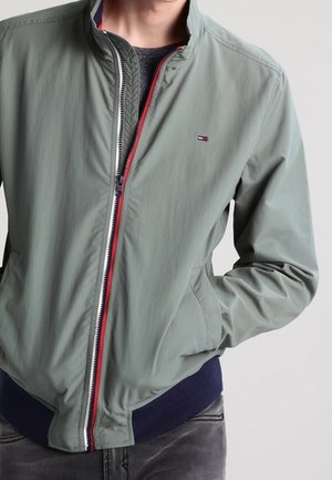 Homme portant une veste gris-vert clair partiellement zippée avec un petit logo de drapeau sur la poitrine, une ceinture bleu foncé et les mains dans les poches.