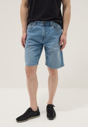 Mann, der hellblaue Jeansshorts, schwarze Sneakers und ein schwarzes Hemd trägt und vor einem einfarbigen Hintergrund steht.