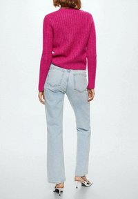 Een vrouw staat met haar rug naar de camera, gekleed in een felroze gebreide trui, lichtblauwe hoge taille jeans en witte sleehakken.