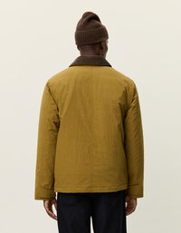 Gul-brun polstret jakke med en mørkebrun krage, senket skuldre og rett snitt. Brukt med en brun strikket lue, vist bakfra.