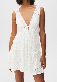 Valkoinen pitsimekko, jossa on syvä V-mallinen kaula-aukko, kukkakuviollisia aukkoja, rento leikkaus ja kerroksittain liehuva helma. Saatavana kevyessä kankaassa.