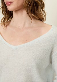 Haut tricoté bleu clair, ample, avec un grand décolleté en V et une texture douce, mettant en valeur une qualité transparente et une apparence légère et aérienne.