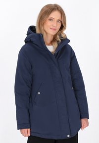 Cămașă impermeabilă de culoare bleumarin, cu glugă, două buzunare laterale, închidere cu capse și o siluetă ușor mai lungă pentru un plus de căldură.