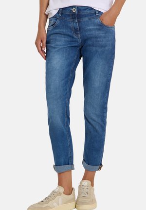 Persona che indossa jeans blu risvoltati e sneaker beige, in piedi con una mano in tasca e l’altra rilassata di lato.