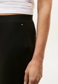Zwarte geribbelde rok met een aansluitende tailleband, met een klein rood, wit en blauw logo aan de zijkant. Glad textuur en strakke pasvorm.