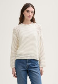 Lys kremfarget strikkegenser med horisontale striper, rund halslinning, løs passform og ribbestrikkede mansjetter, kombinert med blå høyhårede jeans.