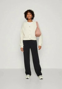 Kremowy sweter z dzianiny o luźnym kroju i z zapięciem na guziki, zestawiony z ciemnymi spodniami typu wide-leg oraz różową torebką na ramię. Sneakersy z akcentami.
