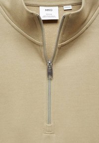 Pull-over couleur beige avec col à fermeture éclair, fabriqué en tissu doux. Doté d'une fermeture éclair argentée et d'une étiquette intérieure indiquant la marque et la taille.