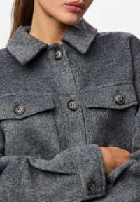 Γκρι μάλλινο σακάκι με λαιμοκόψη με γιακά, δύο τσέπες στήθους και μεγάλα κουμπιά. Απαλή υφή με ελαφρώς βουρτσισμένο φινίρισμα.