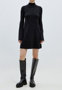Robe noire en tricot avec un col montant et des manches longues, s'évasant légèrement à l'ourlet. Associée à de hautes bottes en cuir noir.